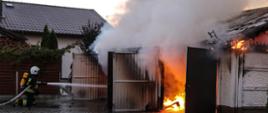 Zdjęcie przedstawia strażaka, który gasi pożar budynku garażowego dwustanowiskowego. Budynek wykonany z płyt betonowych, dach dwuspadowy wykonany z konstrukcji drewnianej pokryty blachą trapezową. Budynek cały w ogniu.