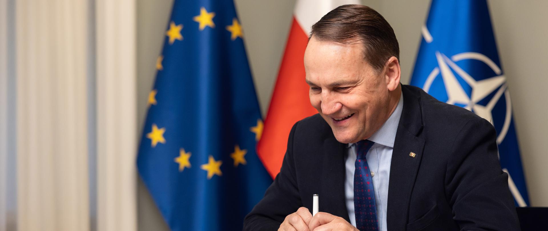 The photo shows a man in a suit sitting at a table. Behind him are three flags: those of the European Union, Poland, and NATO