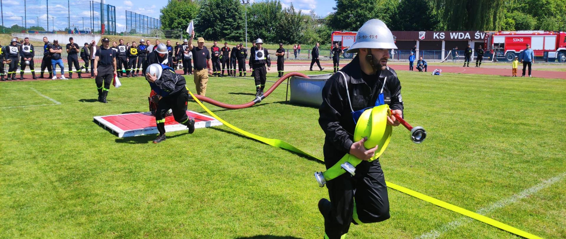 Na murawie boiska druhowie są w trakcie konkurencji ćwiczenie bojowe i biegną z trzymając w rekach węże, w tle widać kibicujących druhów. 