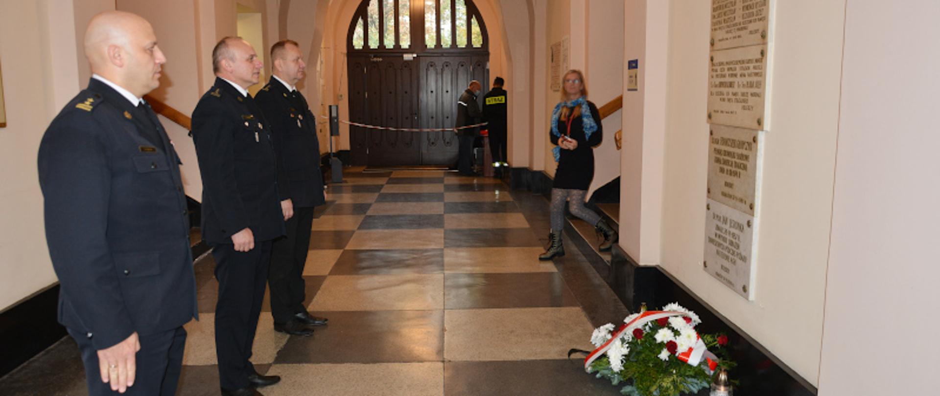 Zdjęcie przedstawia trzech Komendantów Miejskich PSP oddających honory przy tablicy pamiątkowej poległych strażaków w holu Komendy Miejskiej PSP w Krakowie.
