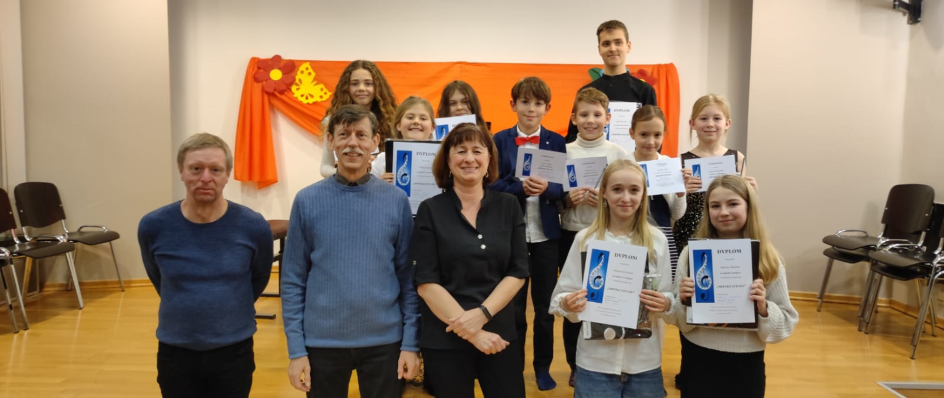 Na zdjęciu grupa osób. Od lewej na dole trzech nauczycieli: dwóch mężczyzn i jedna kobieta. Pierwszy od lewej mężczyzna to blondyn w niebieskim sweterku. Dalej mężczyzna z wąsem i niebieskim sweterku. Na końcu kobieta w brązowych włosach i czarnej bluzce. Koło nich stoją odświętnie ubrani uczniowie, którzy trzymają dyplomy.