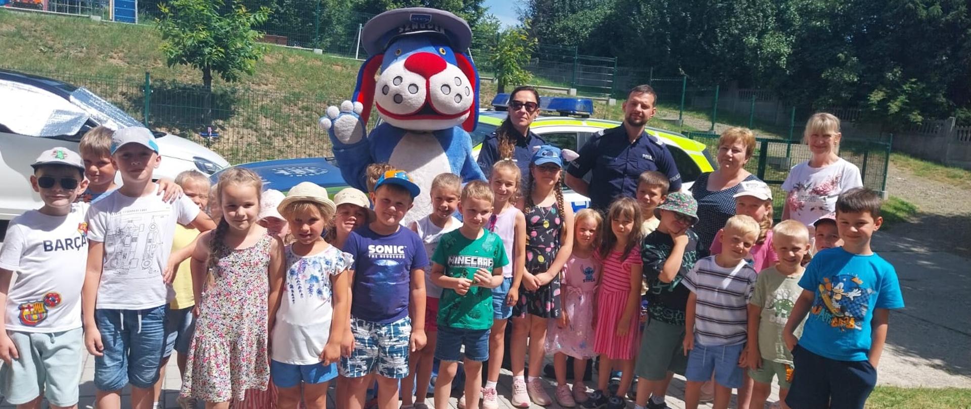 Na placu przy przedszkolu grupa 24 przedszkolaków pozuje do zdjęć w towarzystwie maskotki psa policyjnego Sznupka, dwójki policjantów i dwóch pracownic KRUS.