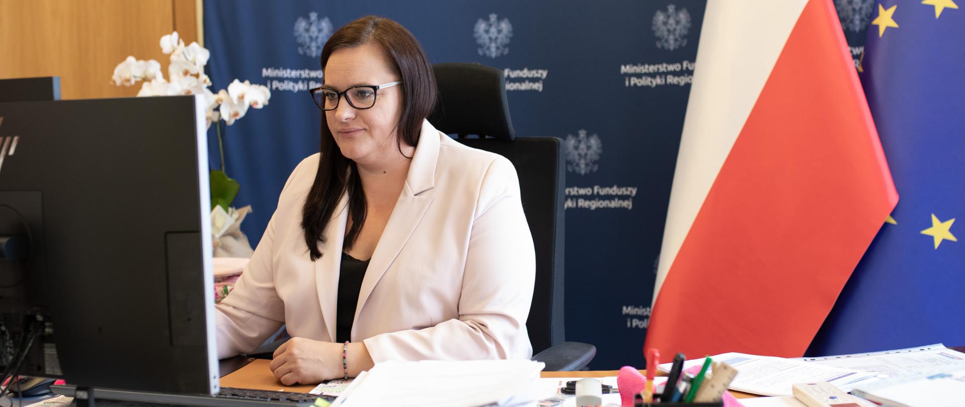 W gabinecie przy biurku przed monitorem siedzi wiceminister Małgorzata Jarosińska-Jedynak. Za nią ścianka reklamowa MFiPR oraz dwie flagi PL i UE.