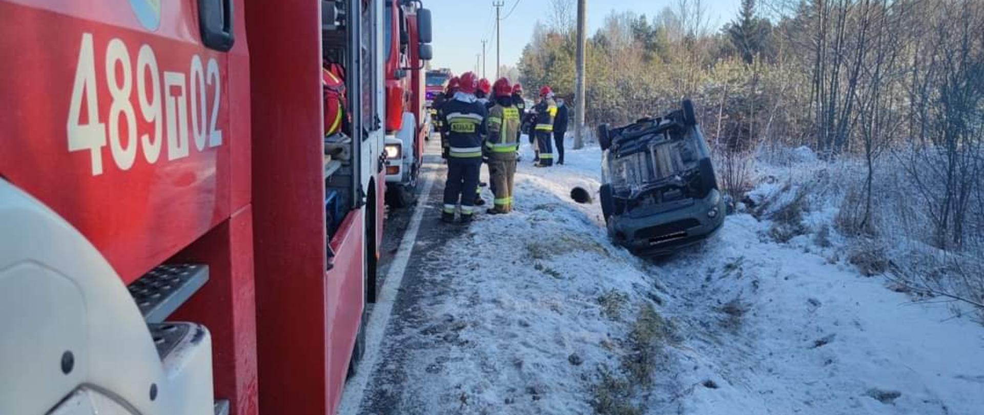 Zdjęcie obrazuje samochód osobowy, który leży na dachu w przydrożnym rowie. Przy pojeździe stoją na poboczu strażacy ubrani w ubrania bojowe oraz samochody pożarnicze z otwartymi skrytkami na sprzęt. Na ziemi leży spora ilość śniegu, na którym widać ślad jaki zrobił pojazd wjeżdżający do rowu