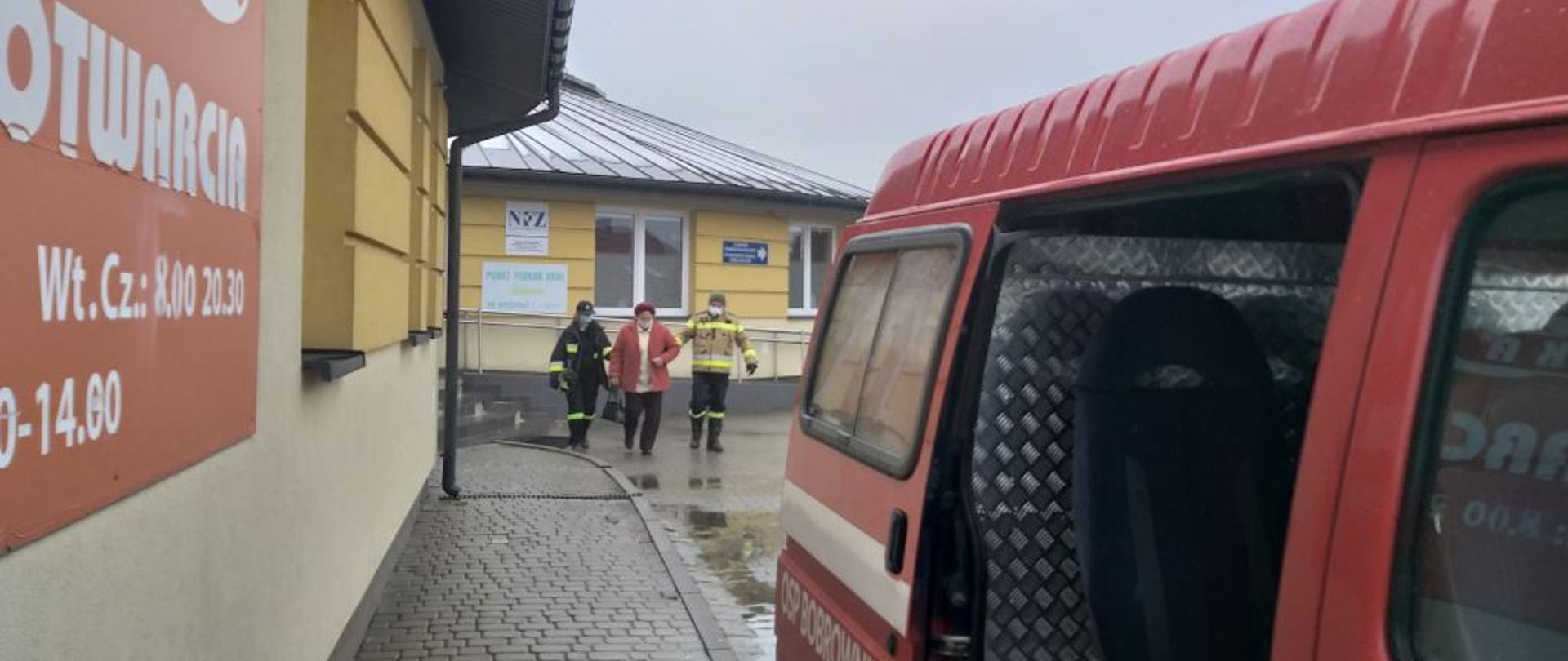 Na zdjęciu na pierwszym planie samochód pożarniczy, którym przewożone są osoby na szczepienia, w oddali przychodnia w której odbywają się szczepienia, oraz osoba szczepiona w asyście strażaków