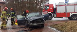 Na zdjęciu w porze dziennej na pierwszym planie widać rozbity przód samochodu osobowego oraz trzech strażaków, którzy prowadzą czynności ratownicze. Na dalszym planie stoi samochód ratowniczo-gaśniczy.