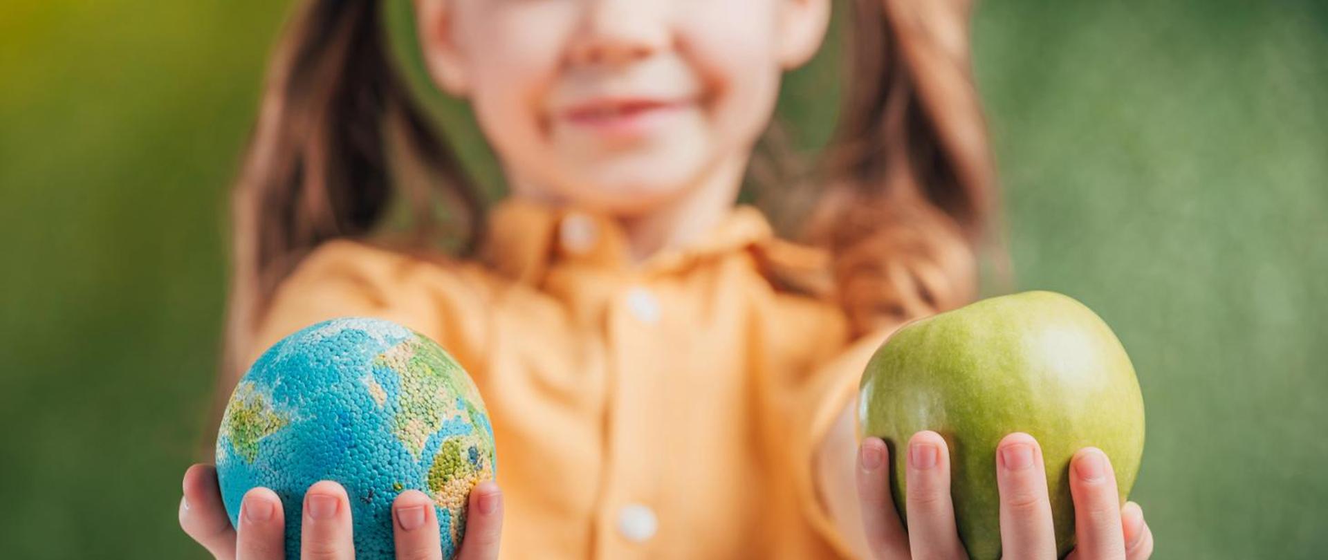 zdjęcie przedstawia małą dziewczynkę, która w jednej dłoni trzyma jabłko a w drugiej mała planetę - ziemię