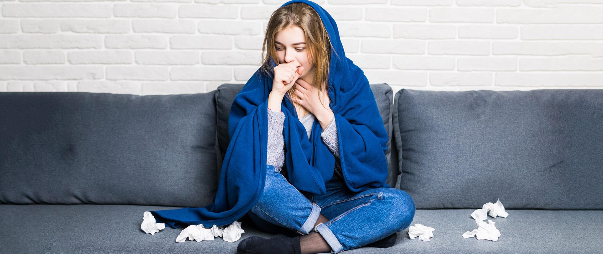 Kobieta w kocu siedzi na kanapie, kaszle. Obok leżą chusteczki higieniczne. 