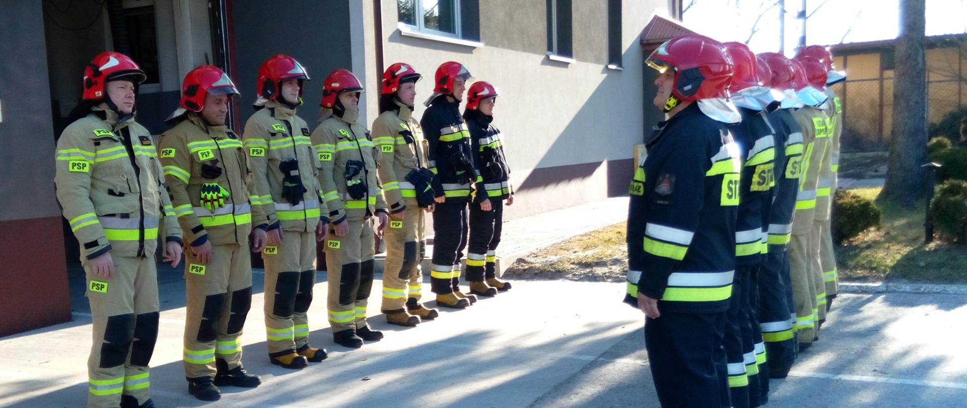 Na zdjęciu przedstawiono zbiórkę zmian służbowych w Jednostce Ratowniczo-Gaśniczej nr 2 PSP w Kielcach. Ratownicy w pełnym umundurowaniu stoją w dwóch szeregach pod budynkiem strażnicy.