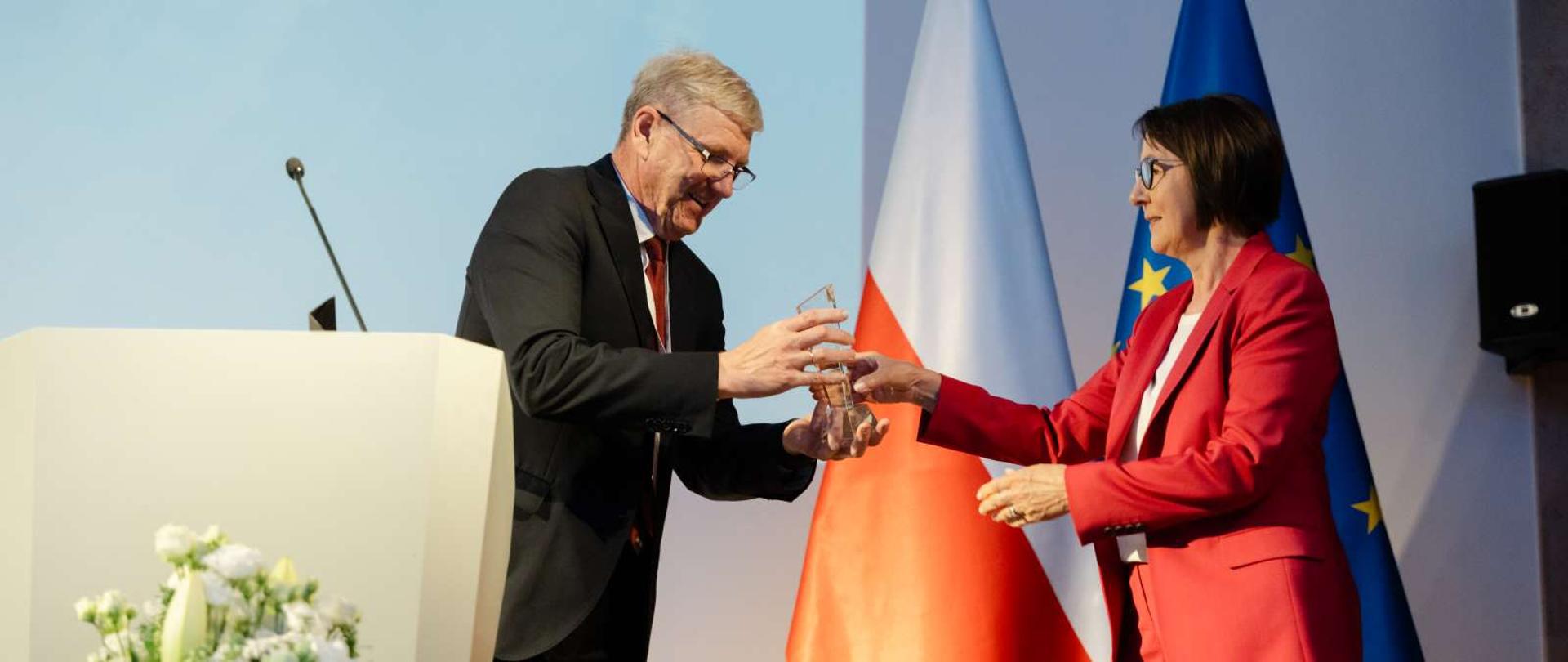 Kobieta i mężczyzna na scenie. W tle flaga biało czerwona, na pierwszym planie białe kwiaty. Mężczyzna wręcza kobiecie statuetkę