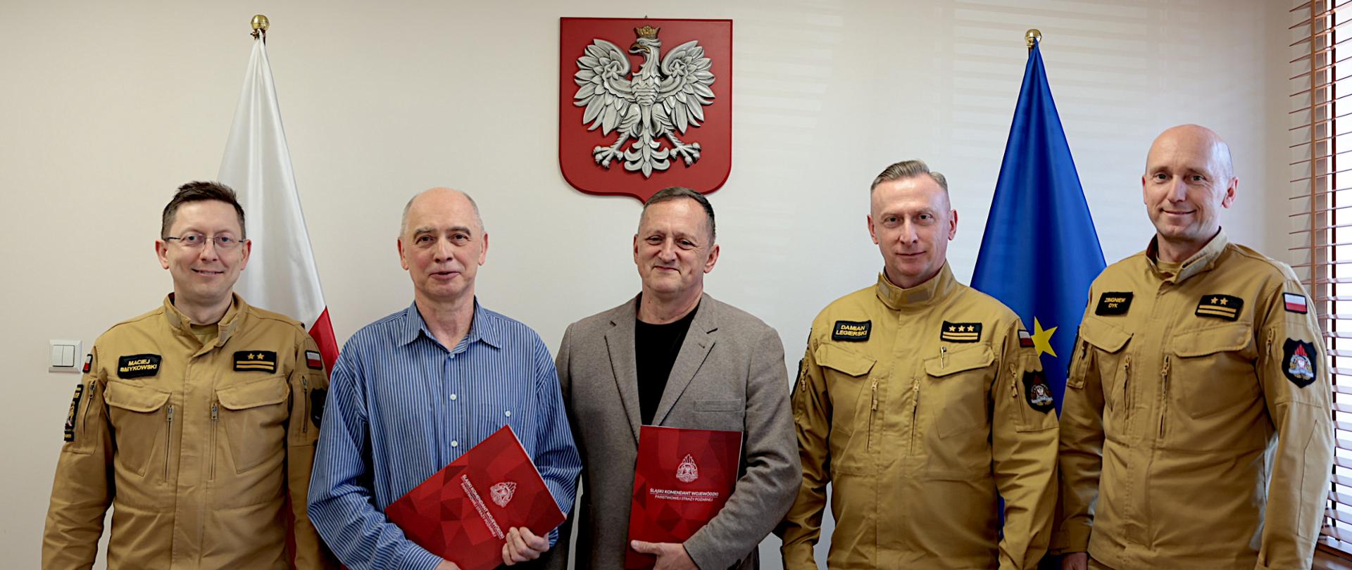 Na zdjęciu widać pięciu mężczyzn stojących obok siebie w jasnym pomieszczeniu biurowym. Trzech z nich, po bokach i z prawej strony, ma na sobie jednolite, piaskowe mundury straży pożarnej. Wśród nich jest starszy brygadier Damian Legierski śląski komendant wojewódzki PSP. Dwóch w środku jest ubranych cywilnie – jeden w niebieską koszulę, drugi w marynarkę. Dwaj mężczyźni w środku trzymają czerwone teczki z dokumentami. Wszyscy stoją przodem do zdjęcia i lekko się uśmiechają. Za nimi na ścianie znajduje się godło Polski, a po obu stronach stoją flagi – polska oraz Unii Europejskiej. 