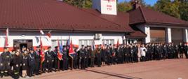 Przed budynkiem remizy Ochotniczej Straży Pożarnej w Jadachach stoją uczestnicy uroczystości. Wśród zgromadzonych stoi osiem pocztów sztandarowych jednostek OSP z terenu miasta i gminy Nowa Dęba. Na budynku widoczne flagi narodowe oraz związku OSP RP. 