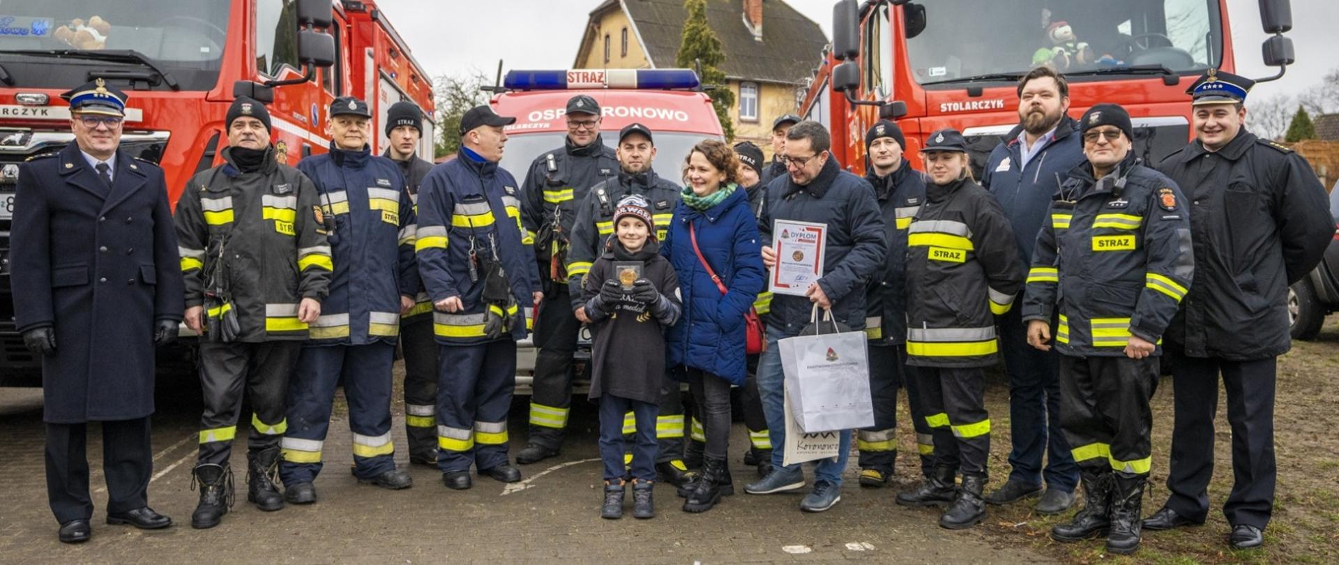 Zdjęcie grupowe, w środku odznaczony chłopiec, przy nim po prawej jego rodzice. Za nimi w szeregu stoją strażacy OSP Koronowo w granatowych ubraniach specjalnych z jasnymi elementami odblaskowymi. W szeregu pierwszy z lewej st. bryg. Jarosław Koprowski, pierwszy z prawej st. kpt. Karol Smarz, trzeci z prawej burmistrz Patryk Mikołajewski. 
