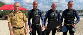 Kolorowa fotografia wykonana na zewnątrz w pogodny dzień na plaży jeziora. Przestawia 3 strażaków płetwonurków ubranych w skafandry nurkowe, jeden ze strażaków ubrany w ubranie służbowe.