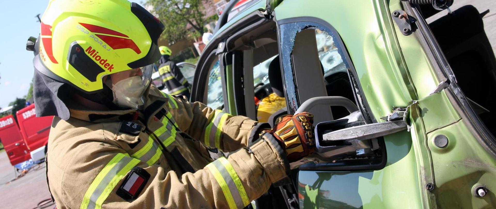 Strażak w żółtym hełmie z napisem "Miodek" i w specjalnym uniformie używa narzędzia hydraulicznego do cięcia zielonego samochodu. W tle można dostrzec inny wóz strażacki oraz kilku strażaków.