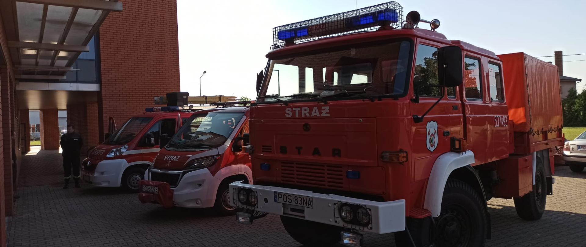 Trzy samochody pożarnicze ustawione przed Komendą Powiatową. Obok nich stoi druh w ubraniu koszarowym.