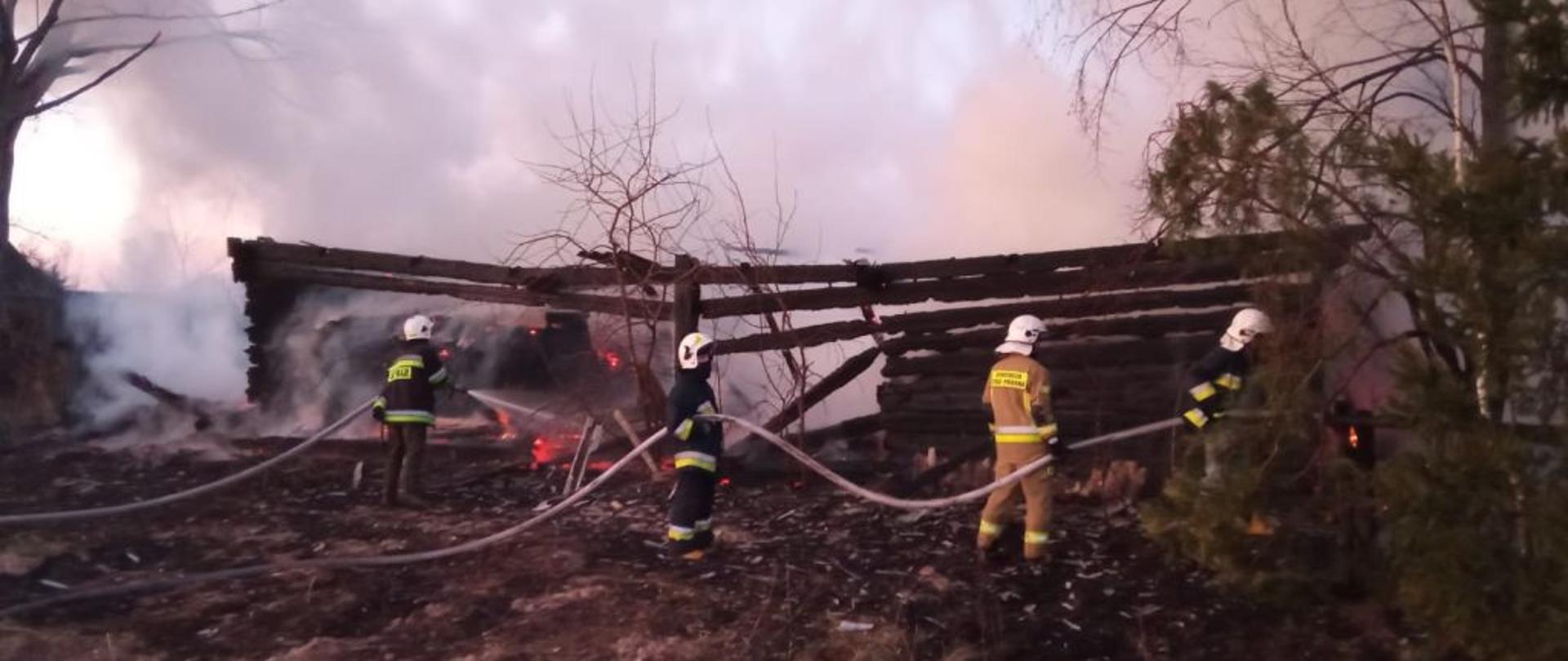 Czterech strażaków w bojowych mundurach bojowych dogasza spaloną konstrukcję drewnianą konstrukcję budynku.