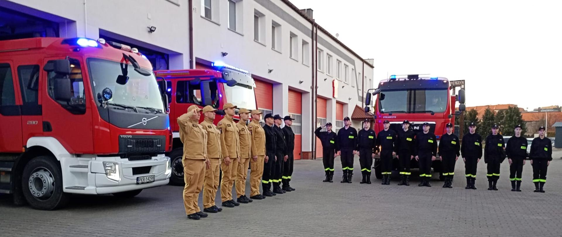 Strażacy stoją w szyku na tle pojazdow pożarniczych usytuowanych na placu komendy, oddając hołd koledze, który zginął podczas ćwiczeń w komorze dymowej