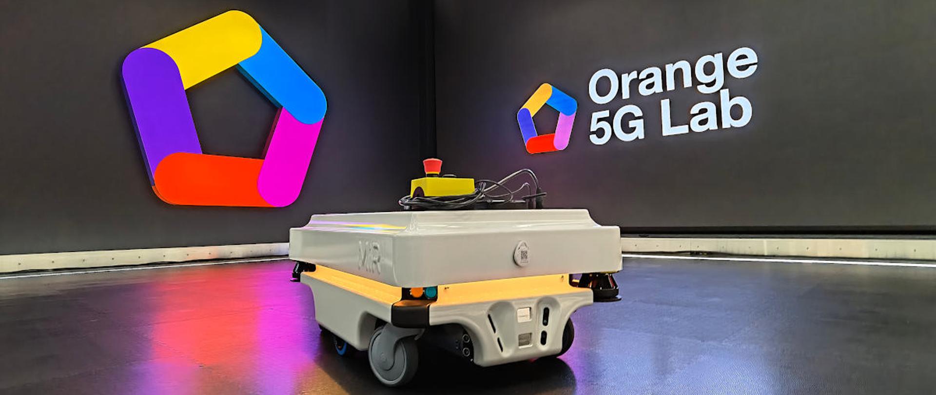 Zdjęcie przedstawia pomieszczenie z umieszczonym na ścianie logo w kształcie pięciokąta w kolorach żółtym, niebieskim, różowym, czerwonym i fioletowym oraz napisem: Orange 5G Lab. Na podłodze pomieszczenia stoi niewielki pojazd w kolorze szarym, kierowany za pośrednictwem sieci 5G.