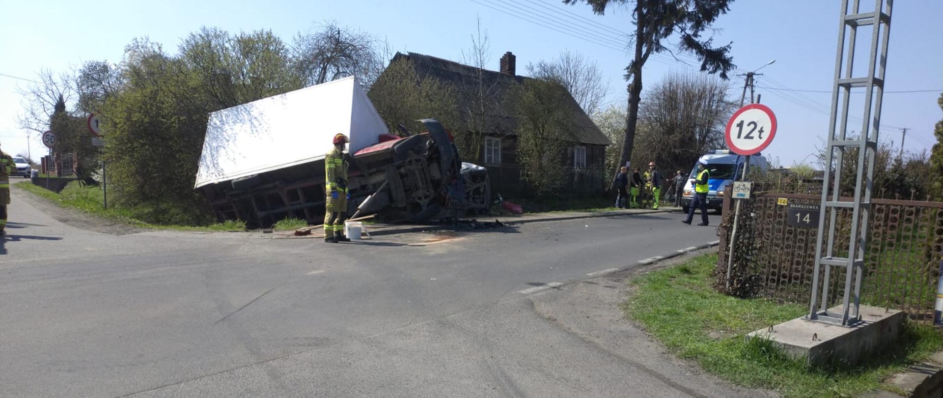 Na zdjęciu widać uszkodzone pojazdy, osobowy i dostawczy. Po prawej stronie od pojazdów opuszczony dom drewniany z metalowym płotem przed. Obok radiowóz policyjny z sygnalizacją ostrzegawczą na dachu. W pobliżu funkcjonariusz Policji, PSP oraz osoby postronne. Prawa strona zdjęcia przedstawia metalowy płot w kolorze brązowym oraz słup i znak drogowy.Po lewej stronie zdjęcia widoczne ogrodzenia posesji z cegły oraz zarośla. W centralnym punkcie funkcjonariusz PSP, za nim szczotka do zamiatania oraz wiadro.