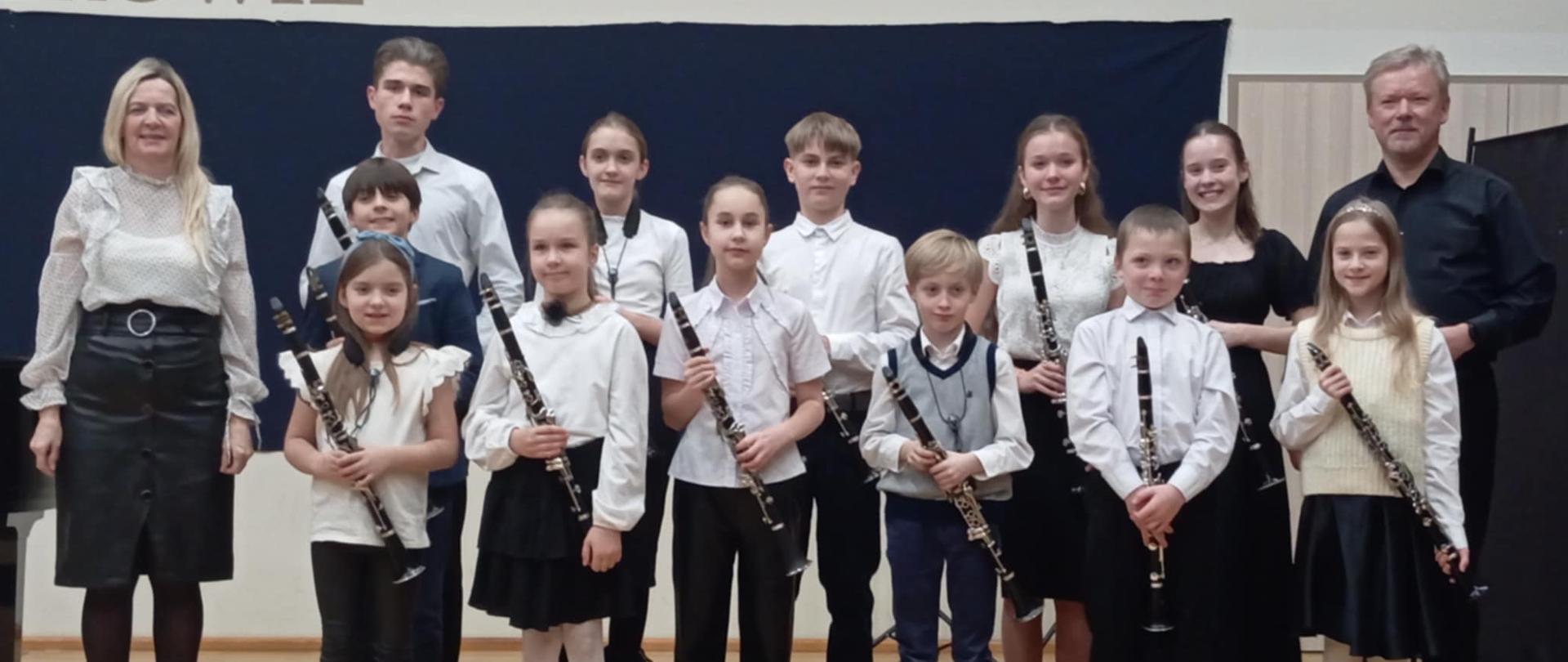 Na estradzie pozuje do zdjęcia w pozycji stojącej 12 uczniów z klarnetami oraz nauczyciele: Anna Łysko i Wojciech Koliński. Zadowolenie i uśmiechy na twarzach klarnecistów są bardzo dobrze widoczne.