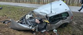 Srebrny samochód w rowie, mocno uszkodzony, za nim strażak