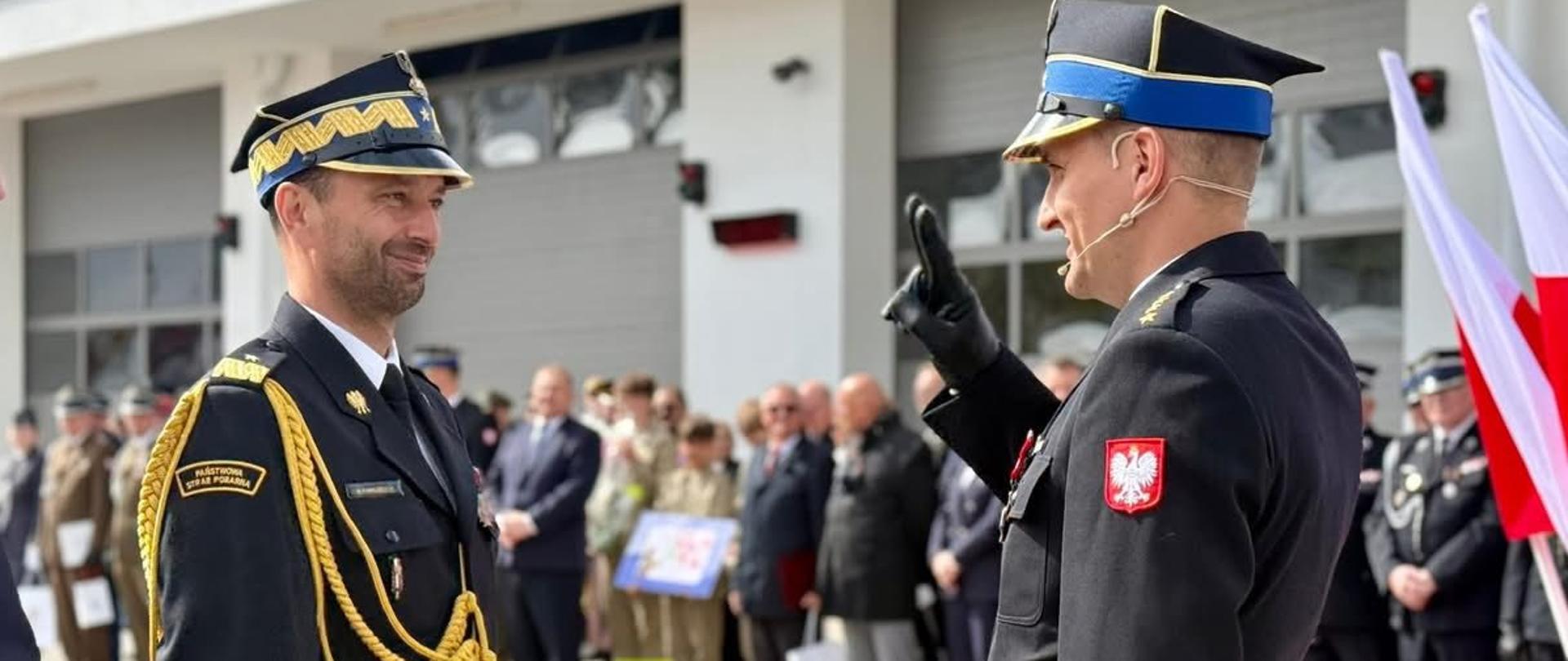 Przed budynkiem strażnicy, na tle dużych bram garażowych, stoją dwaj strażacy w galowych mundurach Państwowej Straży Pożarnej. Jeden z nich unosi rękę w geście salutu. Mundury zdobią sznury galowe i emblematy z orłem. W tle zgromadzeni są inni funkcjonariusze oraz osoby cywilne, a obok widoczne są biało‑czerwone flagi. Scena przedstawia moment oficjalnej uroczystości o patriotycznym charakterze.