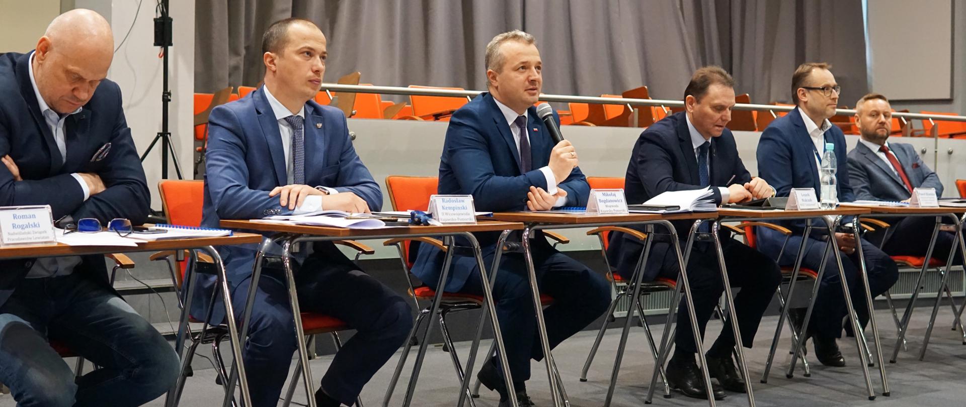 Sześciu mężczyzn siedzi, jeden wypowiada się do mikrofonu