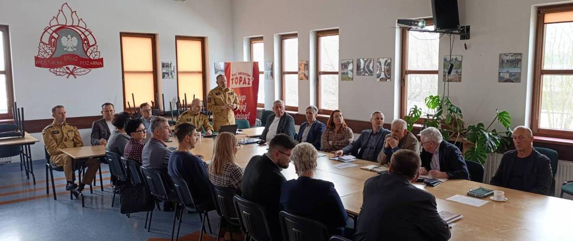 Na zdjęciu widzimy osoby w ubraniach cywilnych przeważają kolory ciemne. Siedzą przy stole. W dalszej części zdjęcia widzimy funkcjonariuszy PSP w ubraniach koszarowych koloru piaskowego. W tle zdjęcia widzimy logo PSP koloru czerwonego na ścianie białego koloru. Na ścianach widzimy okna.