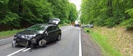 Fotografia przedstawia ujęcie uszkodzonego samochodu osobowego stojącego na środku jezdni, w pojeździe widoczne poważne uszkodzenia lewego przodu oraz lewego przedniego koła, w tle, dalej na jezdni widoczna karetka zespołu ratownictwa medycznego oraz wraki pozostałych dwóch aut biorących udział w zdarzeniu.