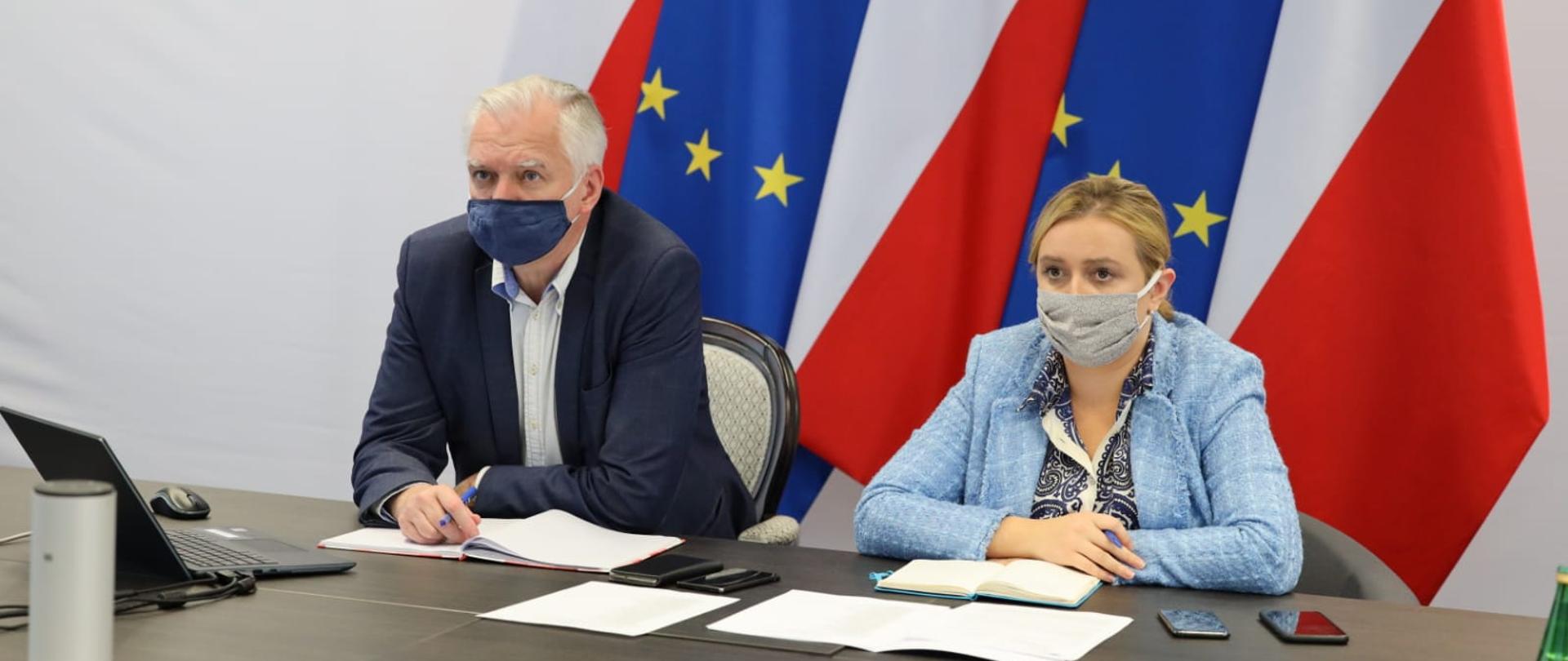 Na zdjęciu wicepremier Jarosław Gowin i wiceminister Olga Semeniuk podczas telekonferencji.
Siedzą za stołem, na stole rozłożone notatki, mają na twarzach maseczki, za ich plecami stoją flagi Polski i Unii Europejskiej.