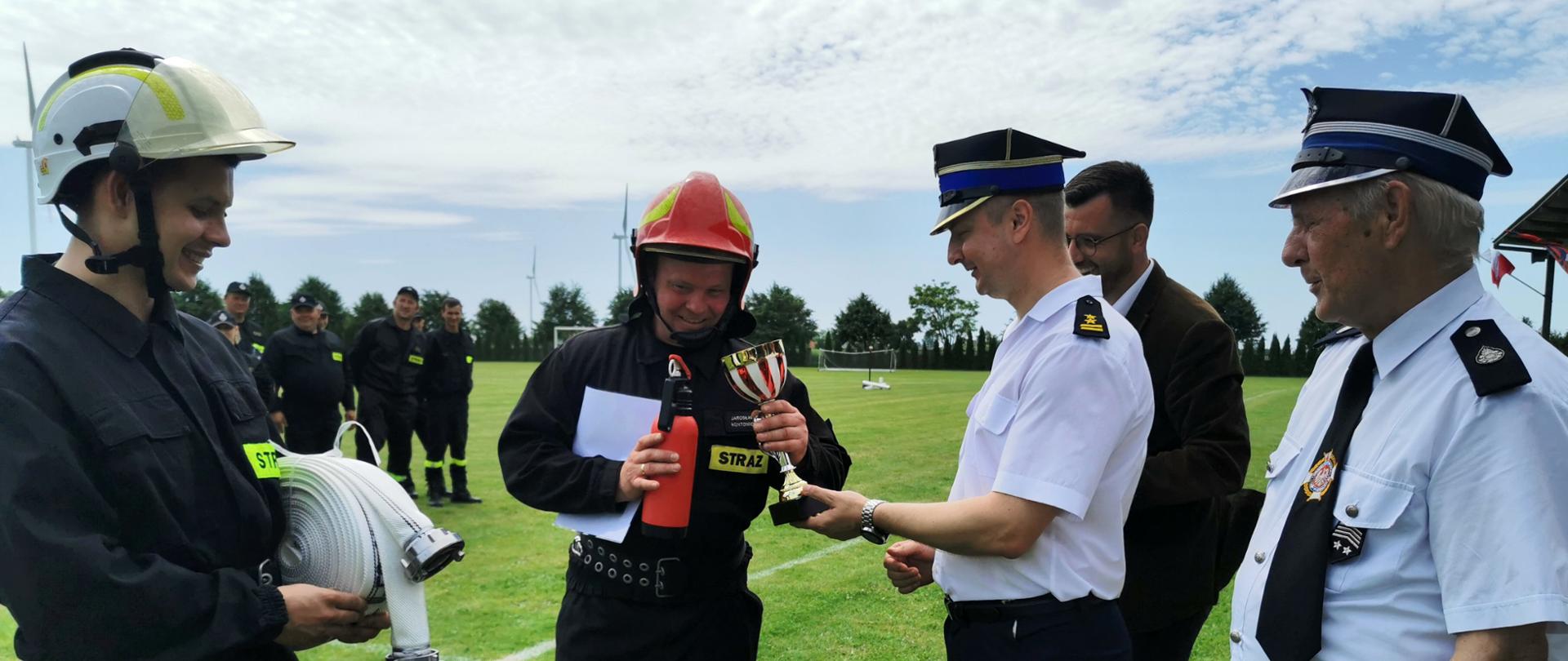 Zdjęcie przedstawia moment wręczania pucharu kierownikowi drużyny zwycięskiej pucharu i pamiątkowych gadżetów. Przy wręczaniu obecni Wójt Gminy Waganiec oraz Prezes Zarządu OSP powiatu aleksandrowskiego