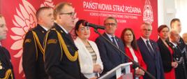 Komendant bryg. Marek Misiura przemawia przy mównicy, Obok niego stoją na tle ścianki z orłem białym, logo PSP oraz napisem Państwowa Straż Pożarna inni strażacy i osoby cywilne.