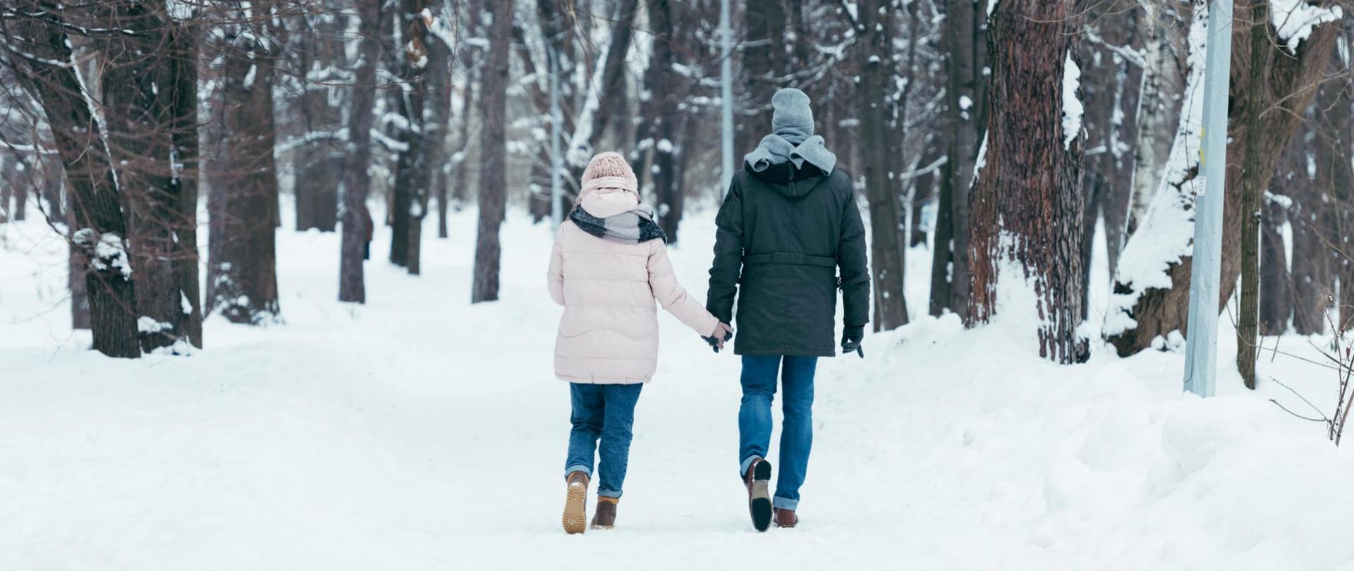 Na zdjęciu są dwie osoby - kobieta i mężczyzna trzymający się za ręce. Znajdują się w zaśnieżonym lesie.