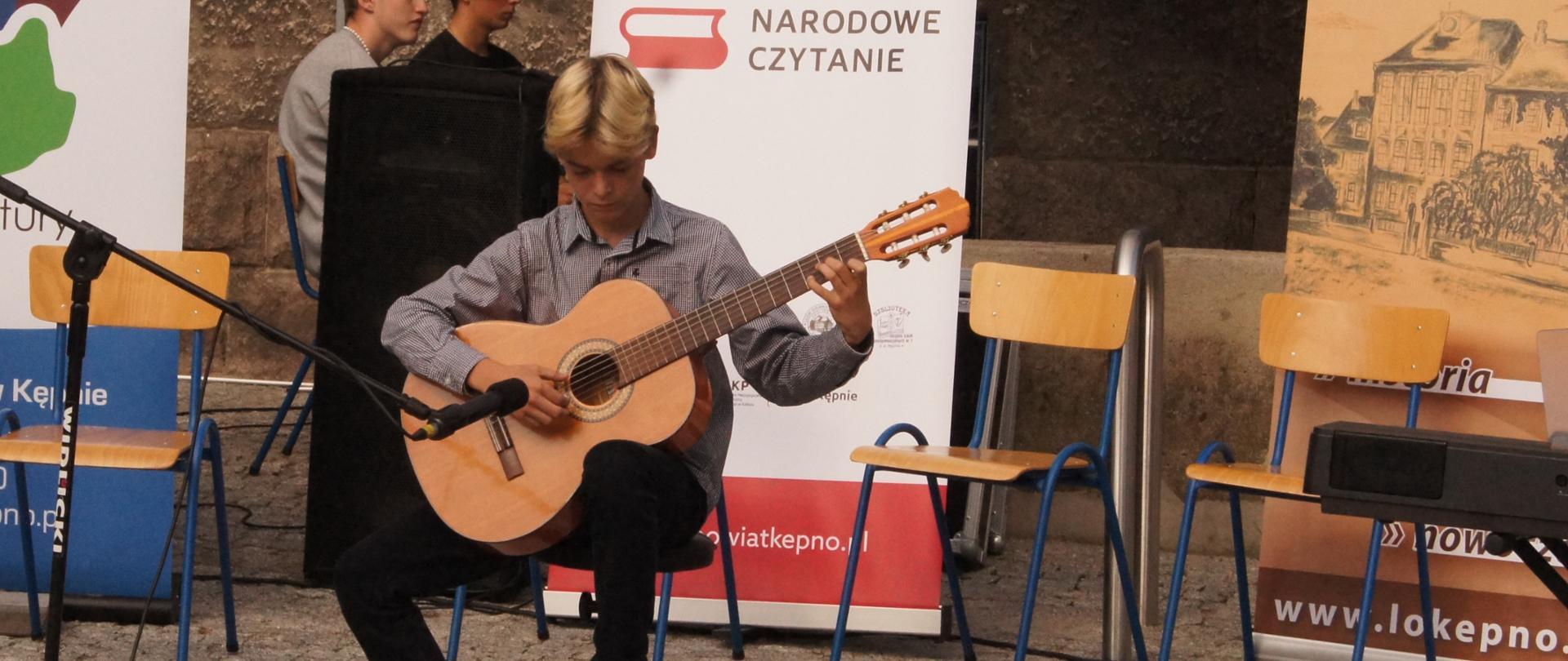 Zdjęcie przedstawia ucznia siedzącego na obrotowym stołku i grającego na gitarze. Uczeń ubrany jest w ciemną koszulę w drobną kratkę, czarne spodnie i buty sportowe. Do gitary przystawiony jest mikrofon. Występ odbywa się na otwartym powietrzu. W tle stoją krzesła szkolne w kolorze jasnego drewna i stojące roll-upy. Od lewej fragment roll-upu powiatu kępińskiego w kolorze biało-niebieskim, od prawej roll-upu LO Nr 1 w Kępnie w kolorach brązowych. Środkowy w barwach narodowych z cytatem "Kto czyta książki żyje podwójnie" Umberto Eco i napisem Narodowe Czytanie. 