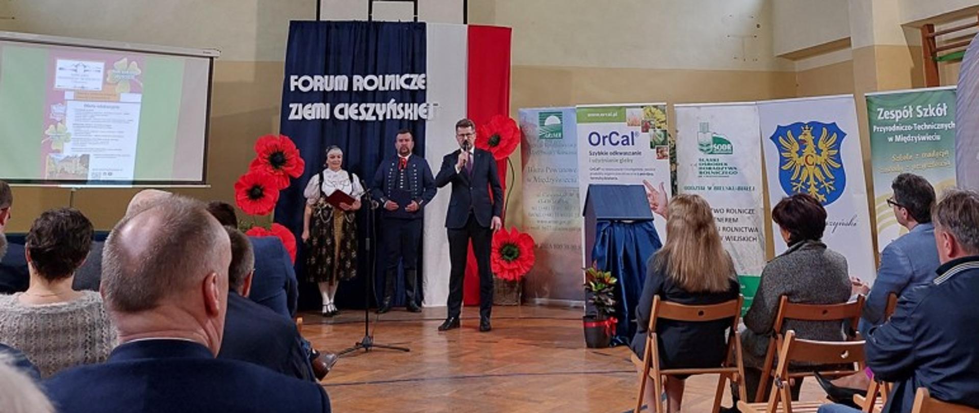 na zdjęciu widok na salę audytoryjną, na scenie prowadzący, witający gości, w tle para ubrana w stroje ludowe, cieszyńskie