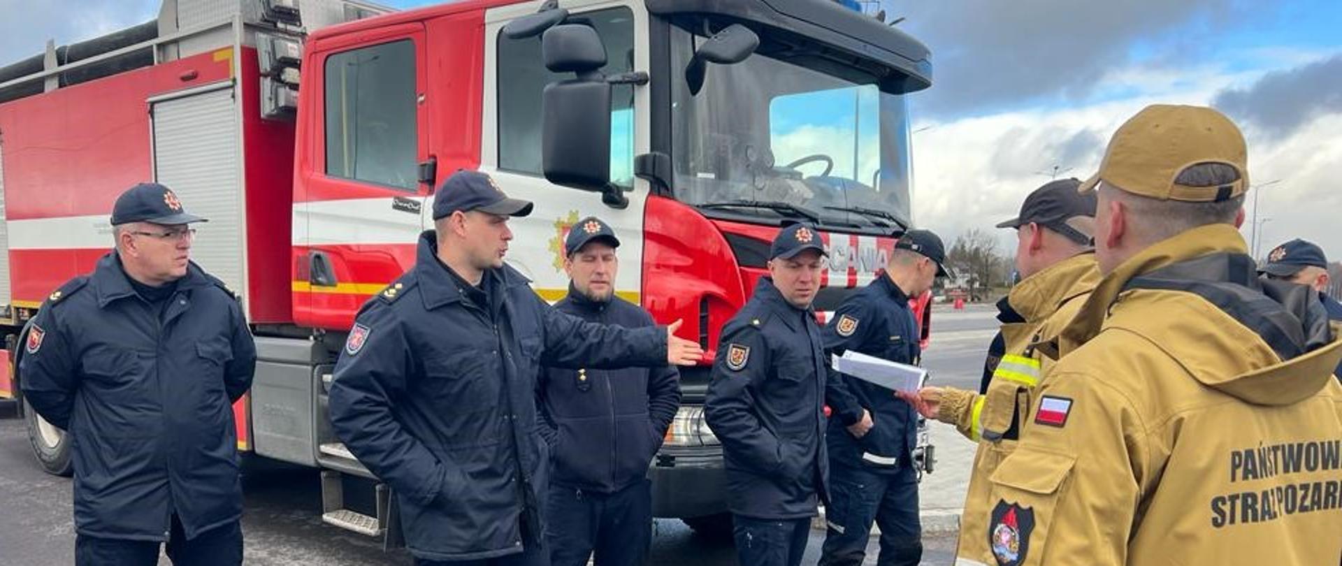 Grupa strażaków z Polski i Litwy stoi przy samochodzie rat-gaś rozmawiając podczas ćwiczeń. Jeden ze strażaków podaje drugiemu dokument