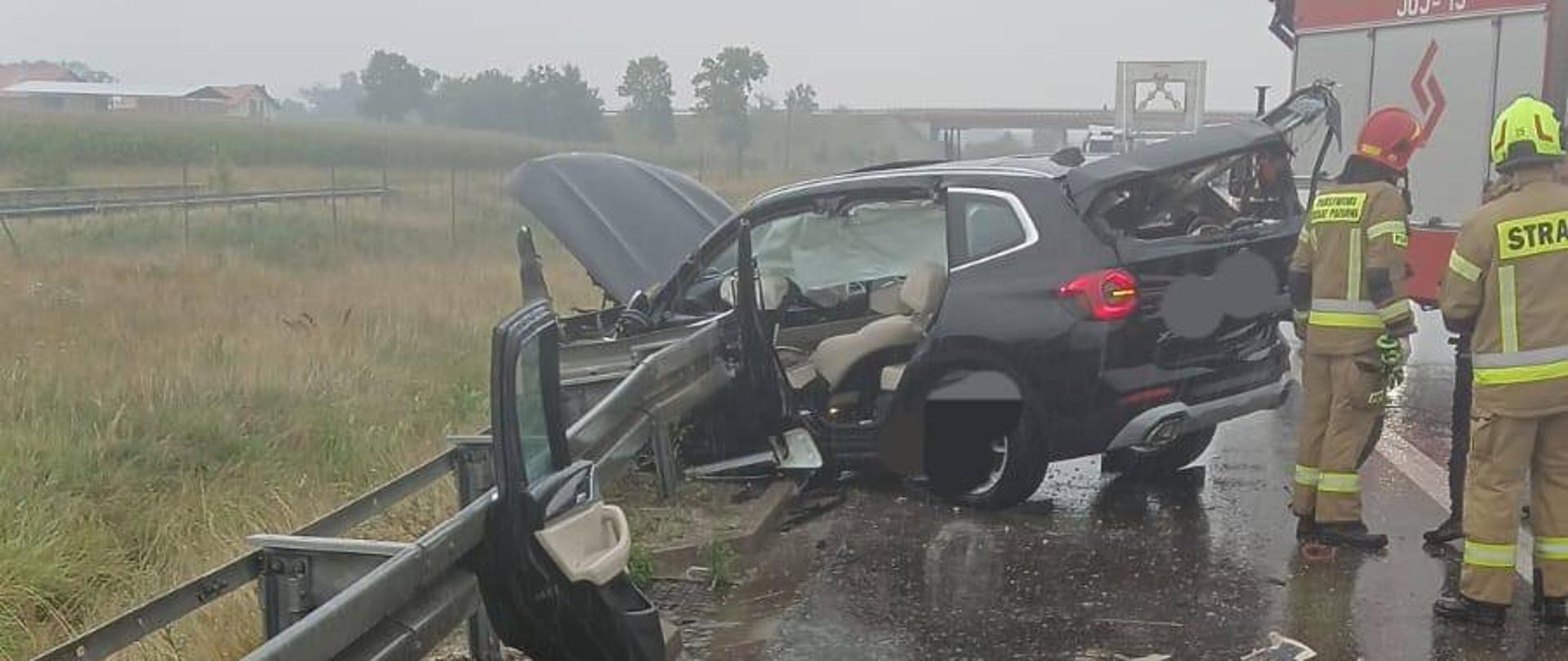 Na zdjęciu widoczna jest scena wypadku drogowego. Czarny SUV uderzył w barierkę energochłonną i zatrzymał się częściowo na niej – przód pojazdu jest poważnie zniszczony, a drzwi kierowcy zostały wyrwane i leżą przy barierce. Auto jest mocno uszkodzone również z tyłu. Na zdjęciu rozrzucone są liczne elementy pojazdu: zderzak, wycieraczka, fragmenty wnętrza aut. W tle widać samochód Straży Pożarnej oraz dwóch strażaków w pełnym umundurowaniu. Warunki pogodowe są deszczowe, jezdnia jest mokra. 