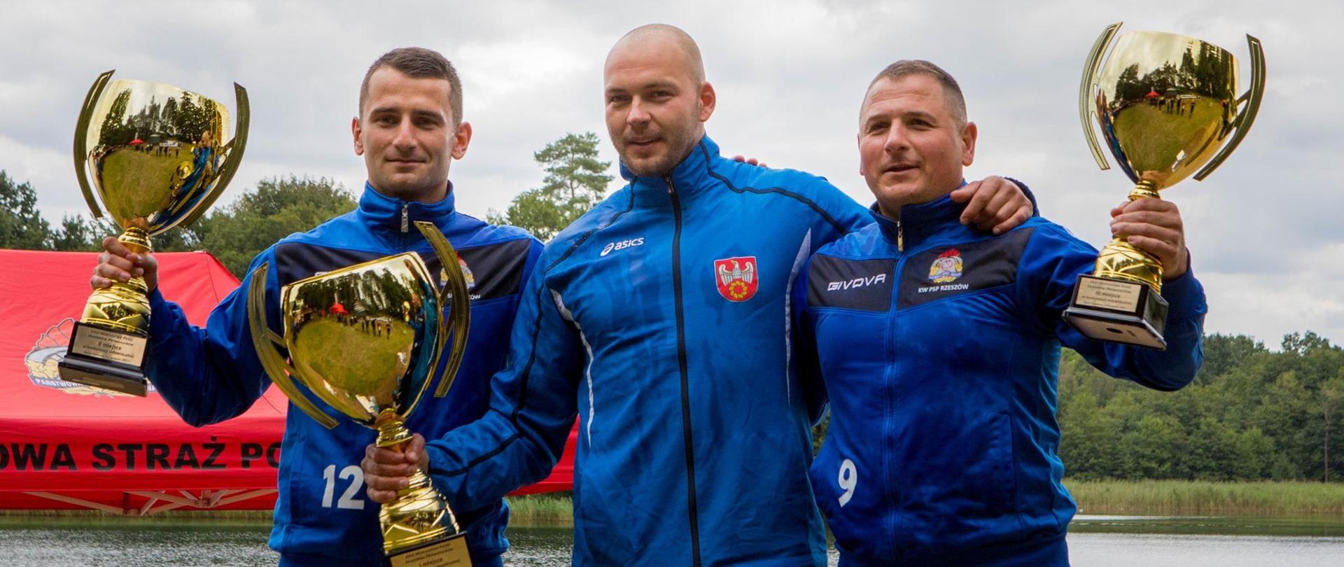 trzech mężczyzn w niebieskich bluzach sportowych i czarnych spodniach trzymający duzy złoty puchar, w tle jezioro