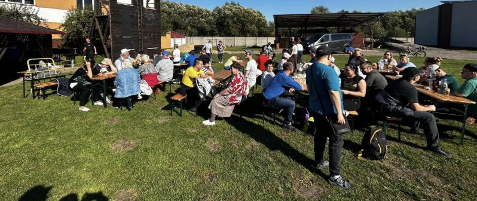 Centralny punkt zdjęcia stanowi grupa ludzi siedząca przy stolikach rozstawionych na terenie zielonym przy budynku Komendy PSP w Białogardzie. W tle widać ścianę wspinali i altankę. Zgromadzeni ludzie jedzą posiłek przy stołach. Jest ładny pogodny dzień.