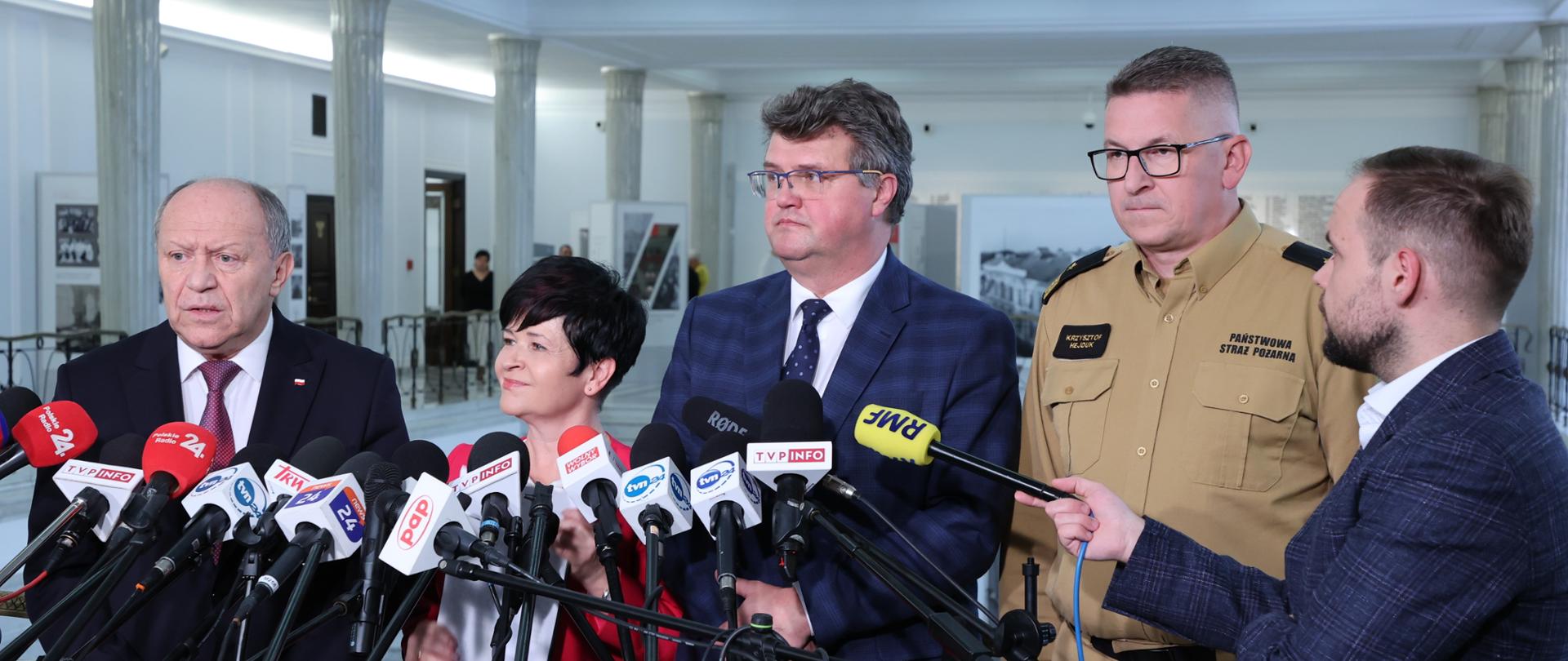 Na zdjęciu widoczni są parlamentarzyści, wiceminister MSWiA oraz Zastępca Komendanta Głównego PSP. Zebrani stoją przed mikrofonami z logami różnych stacji telewizyjnych i radiowych oraz portali internetowych. Odpowiadają na pytania dziennikarzy w sprawie dotacji dla Młodziezowych Drużyn Pożarniczych OSP.