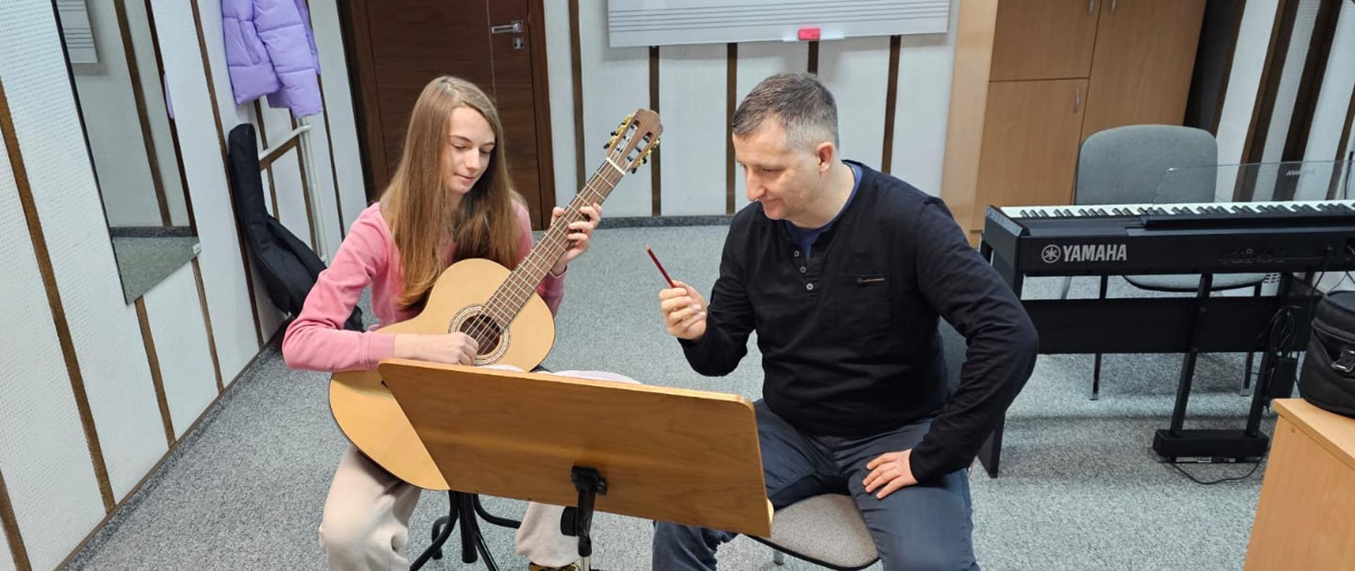 Dziewczyna gra na gitarze, obok siedzi mężczyzna.