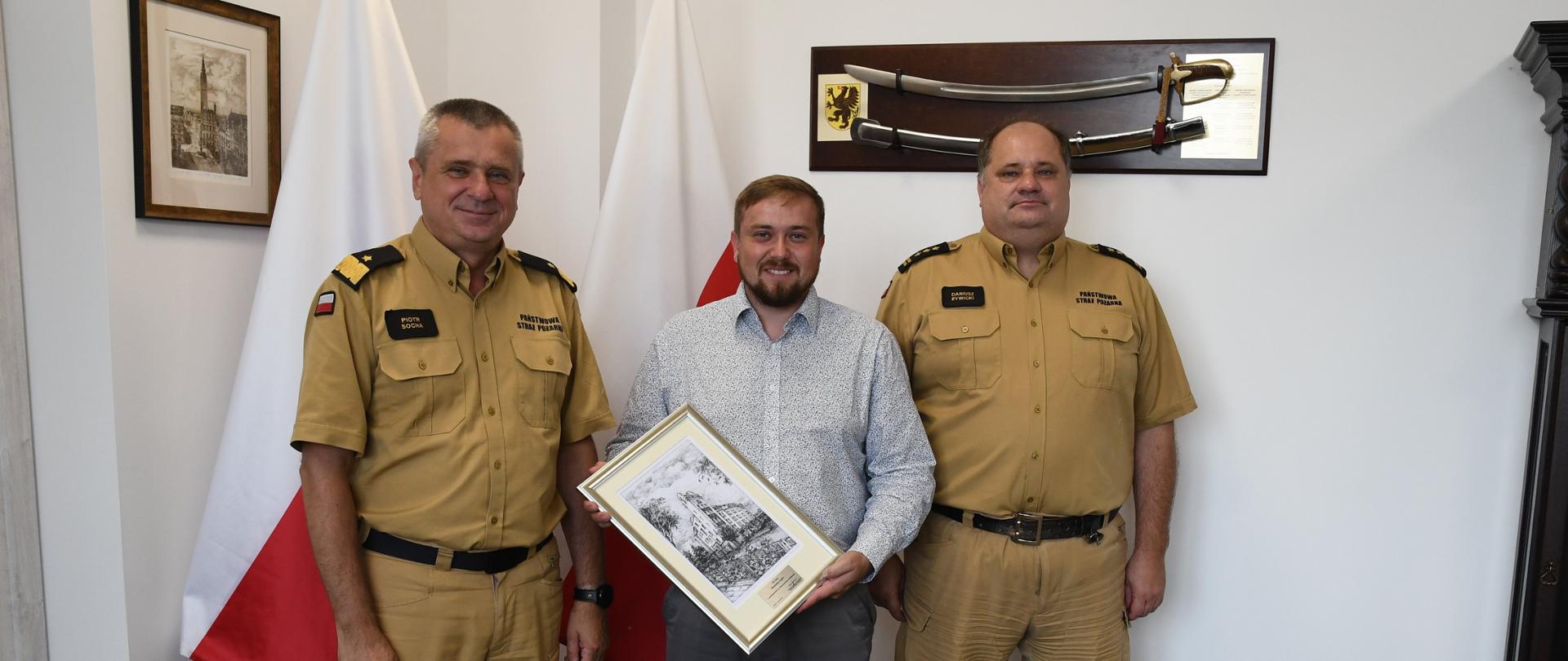 Dwóch strażaków Państwowej Straży Pożarnej stoją z mężczyzną trzymającym obraz za nimi stoją dwie flagi Polski na ścianie wiszą obraz i szabla.