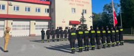 na placu przed Jednostka Ratowniczo Gaśnicza nr 2 stoją strażacy w dwóch szpalerach naprzeciw siebie na uroczystej zbiórce. Pomiędzy szpalerami stoi dowódca. Strażacy ubrani są w czarne ubrania koszarowe. Za szpalerami widoczny jest maszt na który poczet flagowy zawiesza biało-czerwona flagę