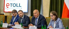 Stół prezydialny przy którym siedzą minister finansów Tadeusz Kościński, szefowa KAS Magdalena Rzeczkowska, pełnomocnik ministra finansów ds. informatyzacji Przemysław Koch, w tle slajd prezentacji z napisem e-Toll.