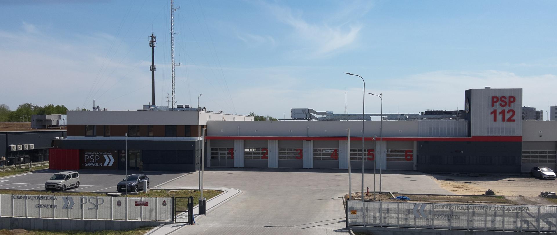 Zdjęcie przedstawia budynek Komendy Powiatowej PSP w Garwolinie w kolorze szaro-czerwonym. Na pierwszym planie ogrodzenie w głębi budynek komendy. W centralnej części bramy garażowe ponumerowane od 1 do 6. Z lewej strony zdjęcia cześć administracyjna komendy. 