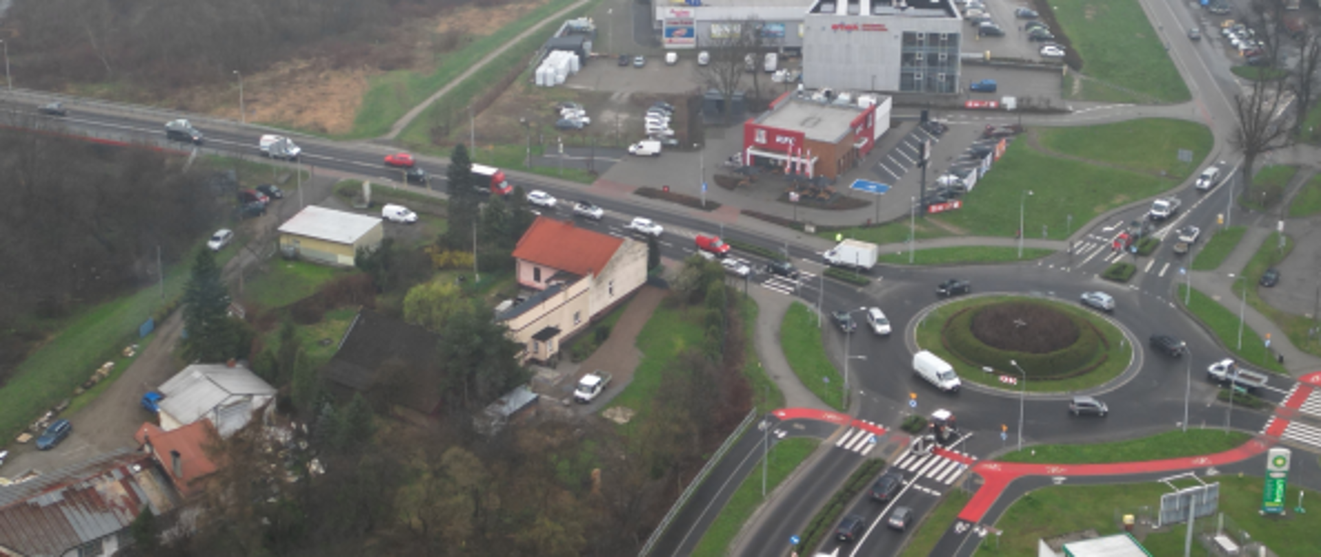 Ujęcie z lotu ptaka przedstawiające skrzyżowanie dróg z rondem w małym mieście, z widocznym ruchem samochodowym, pobliskim centrum handlowym z parkingiem oraz zabudową jednorodzinną; w tle widoczna rzeka i tereny zielone spowite lekką mgłą.