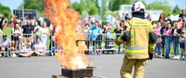 Strażak ubrany w ubranie specjalne trzyma drewniany kij na końcu którego, znajduję się pojemnik. Przed nim leży misa stalowa postawiona na dwóch paletach drewnianych z której unoszą się wysokie płomienie. W tle za barierkami stoją ludzie. 