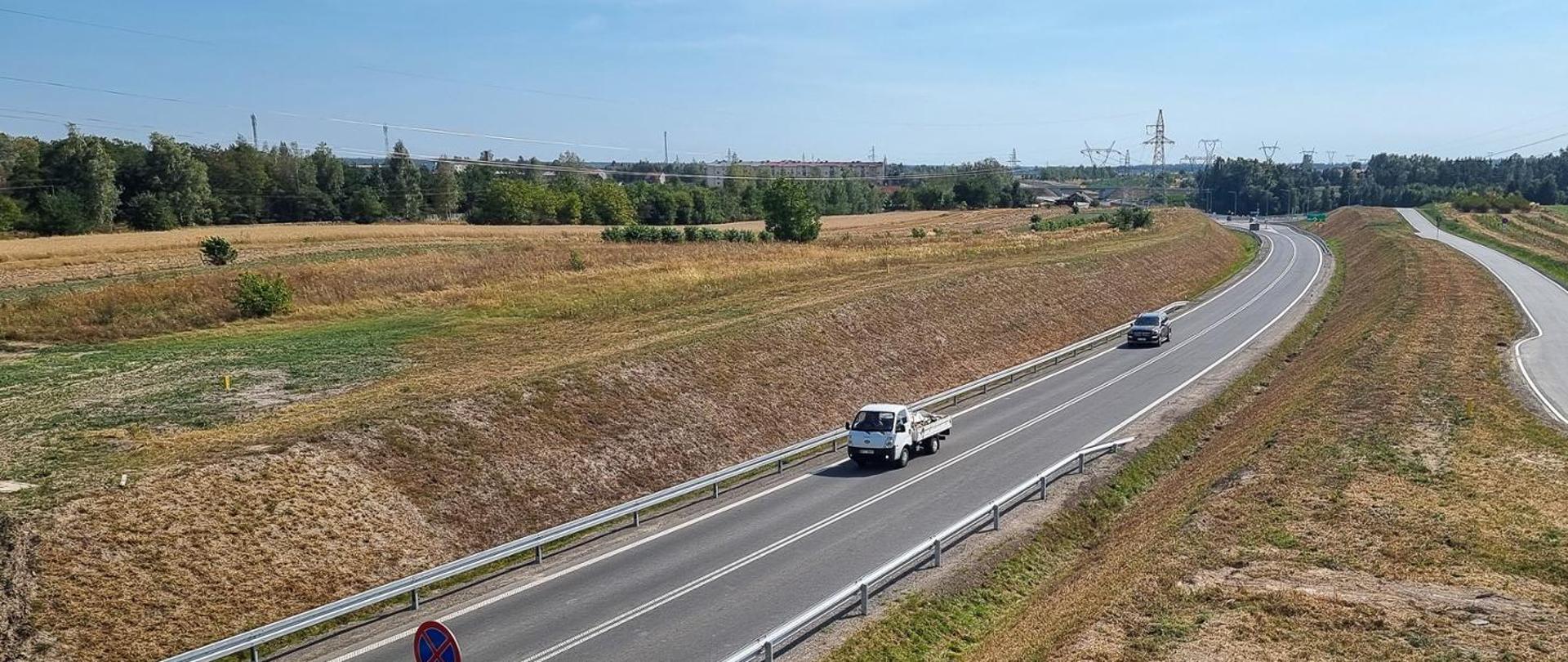 Na zdjęciu widoczna jest spokojna, wiejska sceneria z dwupasmową drogą przecinającą zielone tereny. Po drodze poruszają się dwa pojazdy — biały samochód ciężarowy oraz czarny samochód osobowy. Po lewej stronie drogi znajduje się znak zakazu parkowania. W tle rozciągają się pola porośnięte trawą, pojedyncze drzewa oraz linie energetyczne. Nad całością rozpościera się bezchmurne, błękitne niebo, podkreślające pogodny charakter dnia.