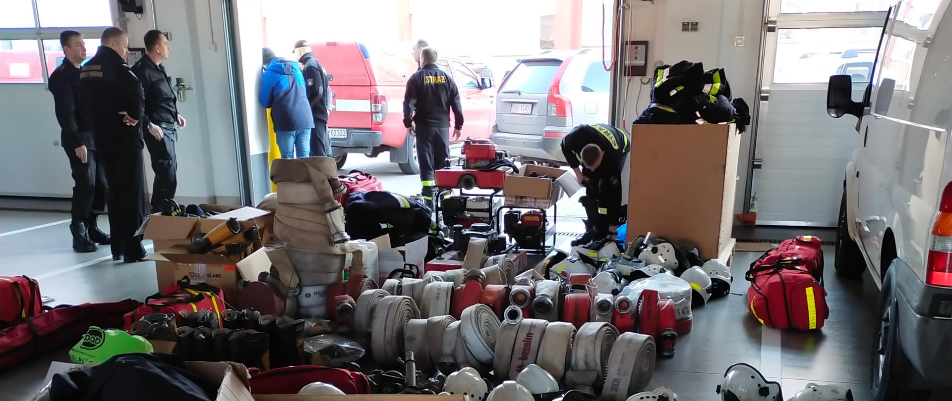 Na garażu Jednostki Ratowniczo-Gaśniczej leży sprzęt pożarniczy. W tle widać funkcjonariuszy przyjmujących go.